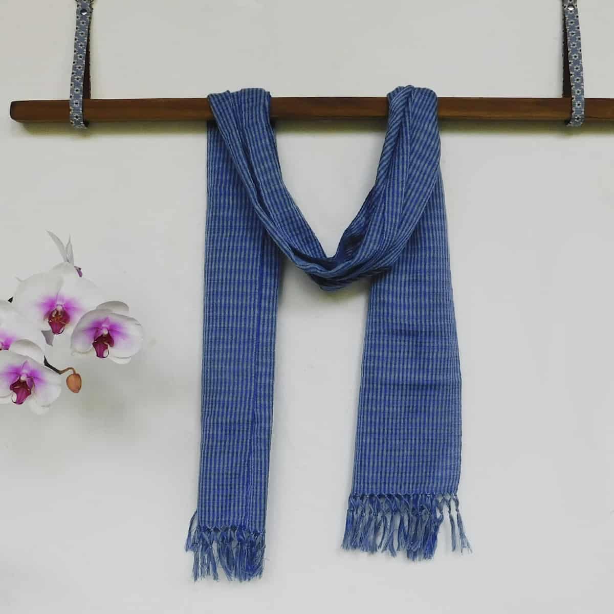Blue and gray scarf hand woven in Guatemala draped over a hanging wooden beam next to white and purple lily flowers