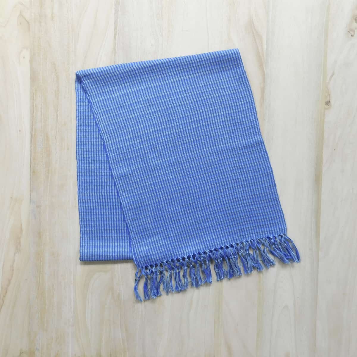 A blue and gray handwoven scarf with a decorative fringe on a light wood background