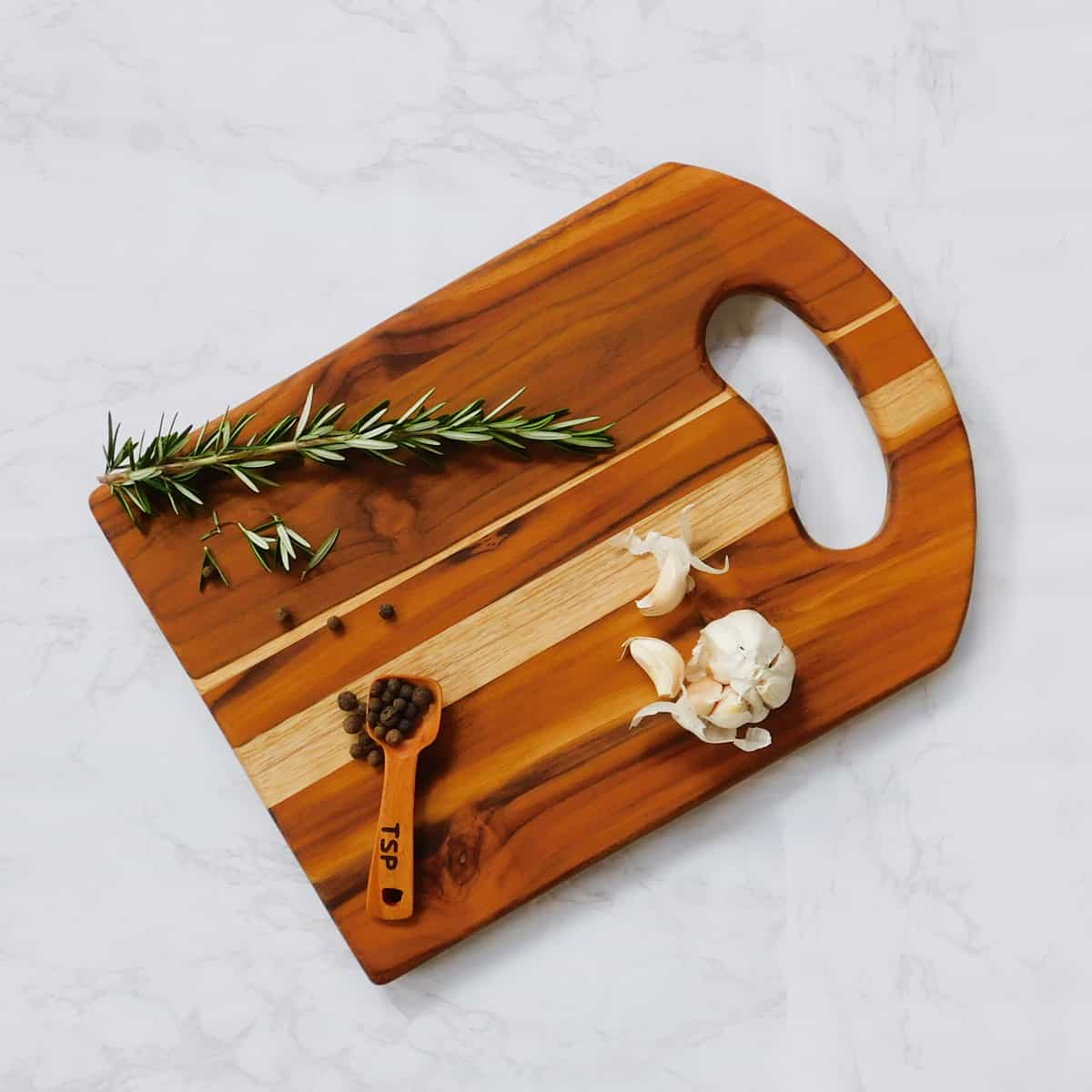 small teak cutting board with rosemary garlic and measuring spoon on white marble background