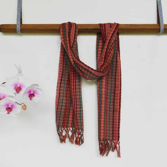 A handwoven earth tones scarf with a decorative fringe draped over a wooden beam next to white and purple lilies.