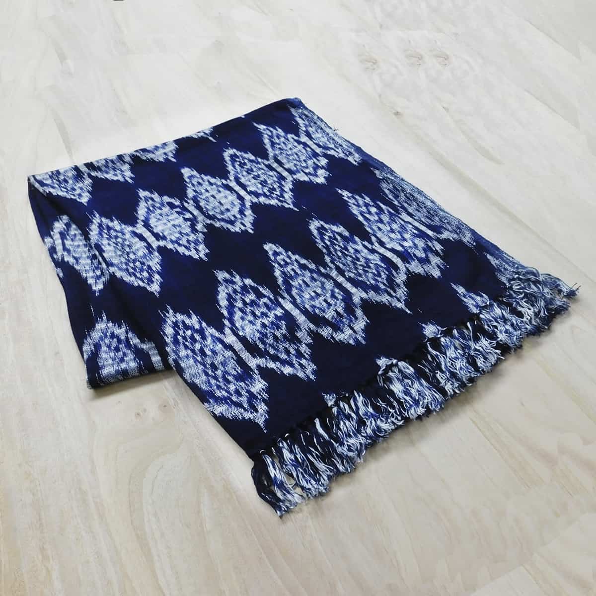 A Guatemalan scarf with a traditional ikat pattern in navy blue and white with a fringe is on a light wood background