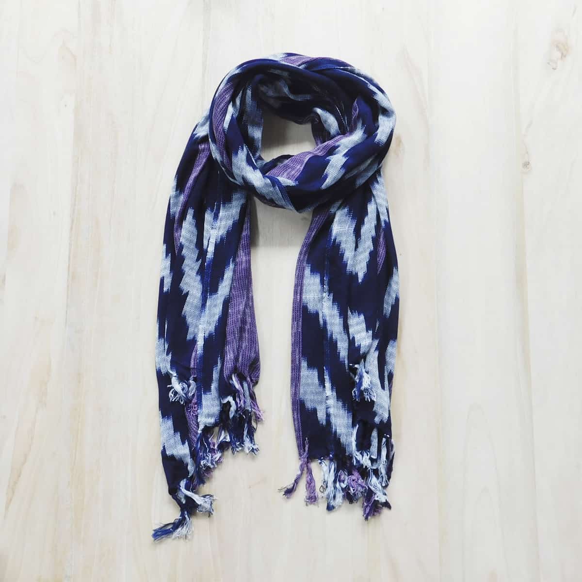 a handwoven guatemalan scarf with navy blue, white and purple stripes in a loop form against a light wood background