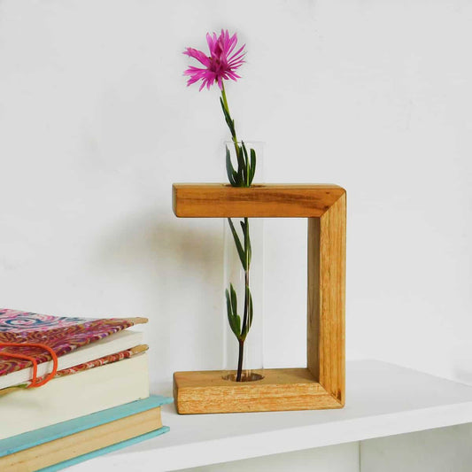 a test tube vase with a pink flower is supported in a three-sided wood frame next to books on a white shelf