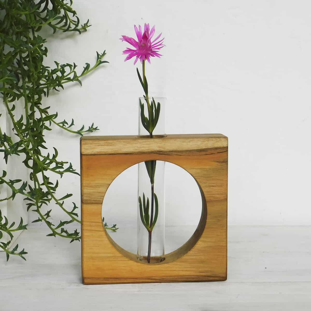  glass tube flower vase with a pink flower supported by a square wooden frame with a circular cutout