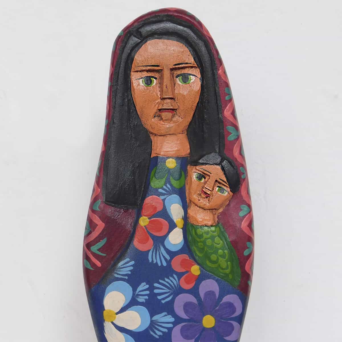 A close-up of a handmade and handpainted virgin mary figurine made of pine wood against a white background. She is holding Jesus and her robes have flowers.