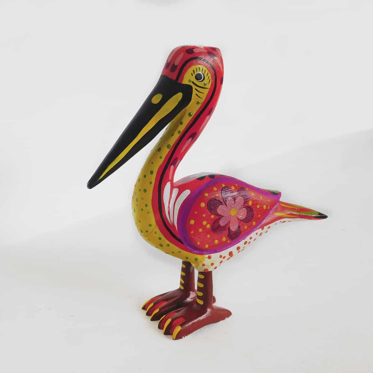 a wooden bird figurine in the shape of a pelican with pink coloring on its back and yellow along its front. its wing features a floral motif.