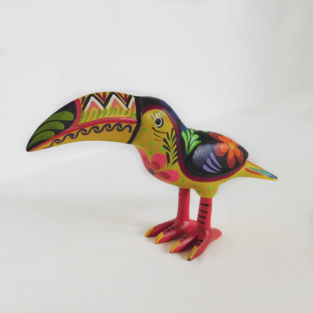 a hand carved wooden figurine in the shape of a toucan with whimsical colors including yellow, purple, pink and orange. its wing has a flower.