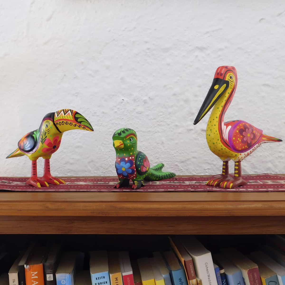 a trio of wild bird figurines on a wooden shelf with a red table runner set against a white wall. the birds are painted in vivid colors.