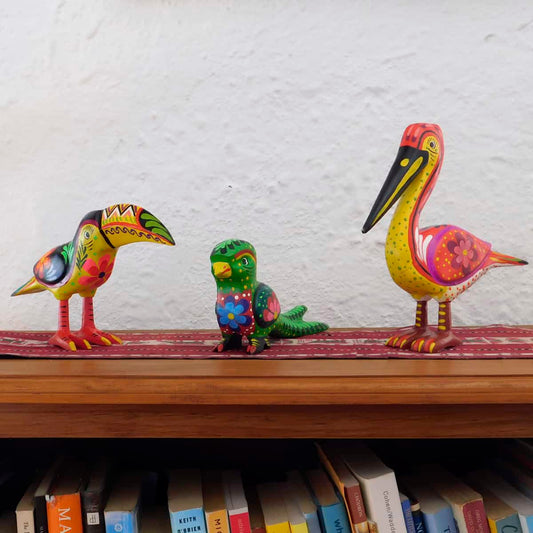 a trio of wild bird figurines on a wooden shelf with a red table runner set against a white wall. the birds are painted in vivid colors.