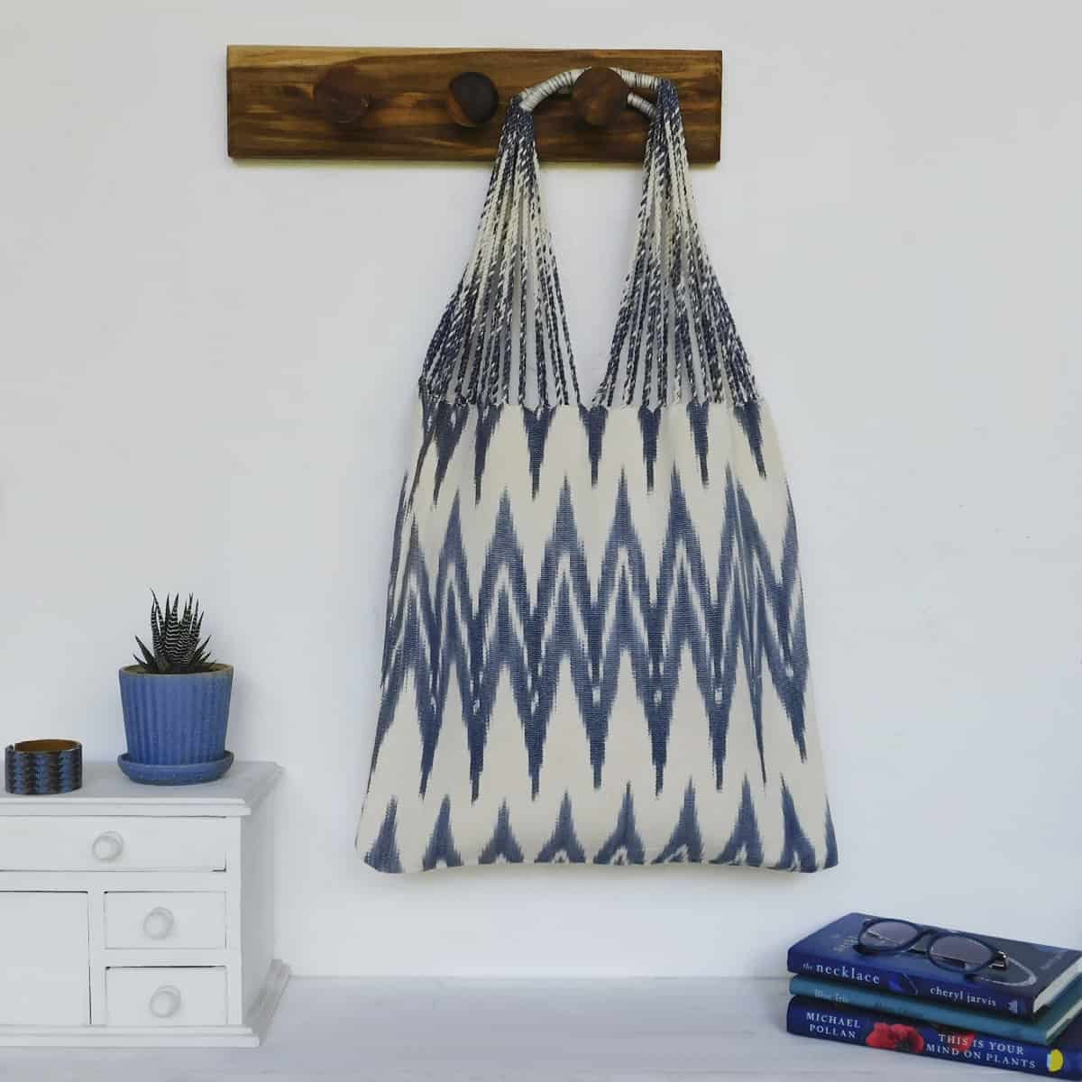 sustainable woven bag in blue gray and off-white in an ikat design woven by Maya weavers hanging next to books and a succulent plant